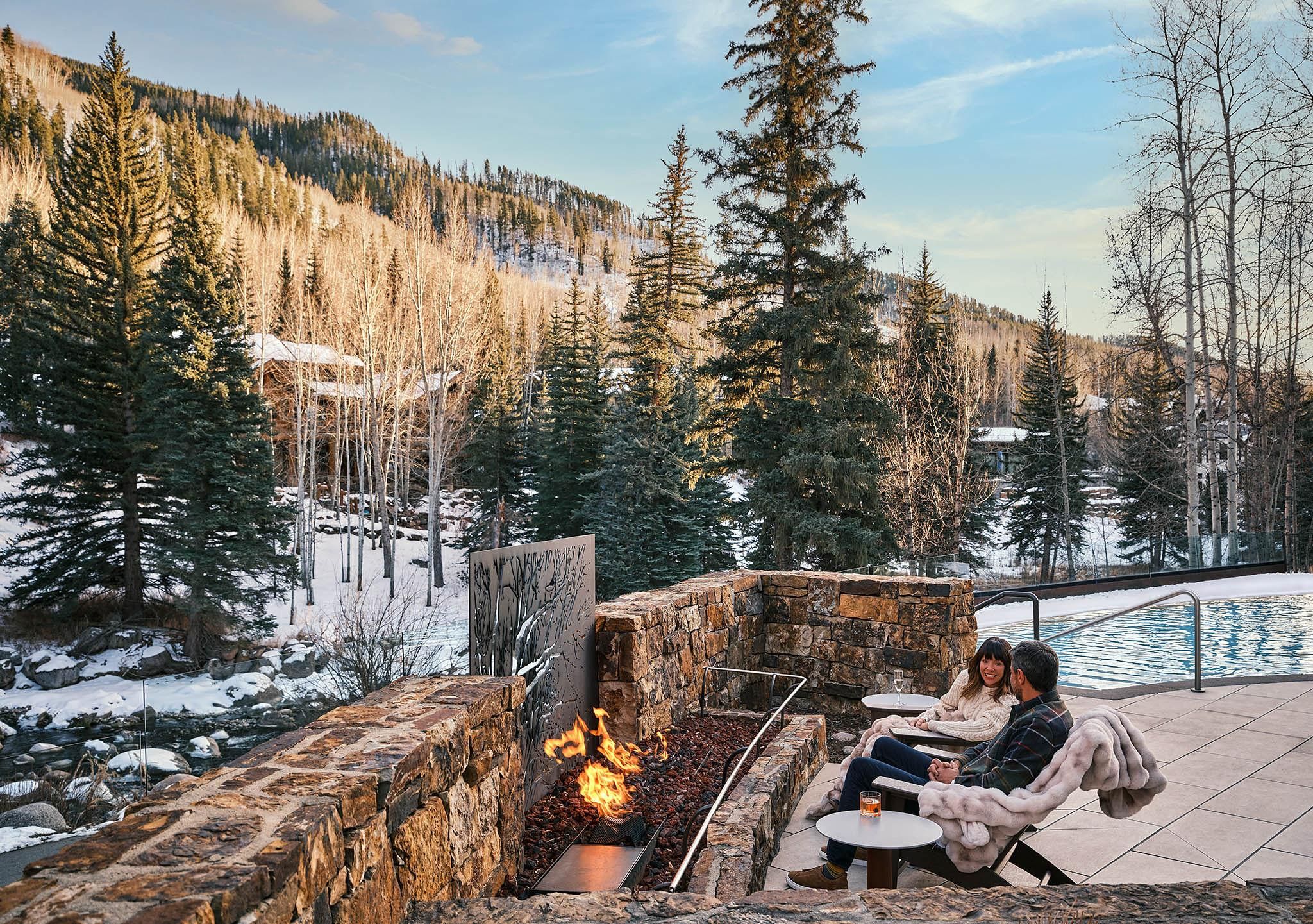 A stone fire pit with a metal grate, surrounded by a stone wall, overlooking a snowy landscape with trees and a clear sky.