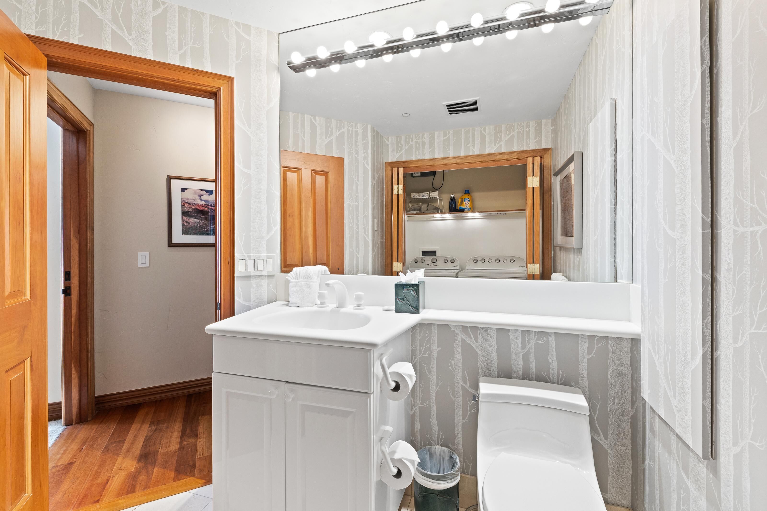 A bathroom with a white vanity, a mirror, and a toilet. There is a wooden door leading to another room, and a trash can is visible.