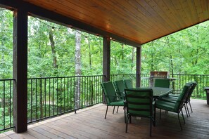 A covered outdoor deck with a wooden table and chairs, surrounded by a metal railing, overlooking a forested area. - Spacious 3600 sq ft 5 king bedrooms with extra bunkbeds (Broken Bow)