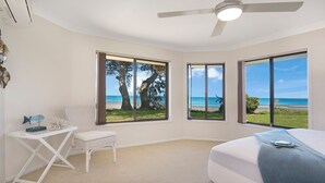 Ein Schlafzimmer mit Bett, Sessel und Schreibtisch. Drei große Fenster bieten Blick auf Strand und Bäume.