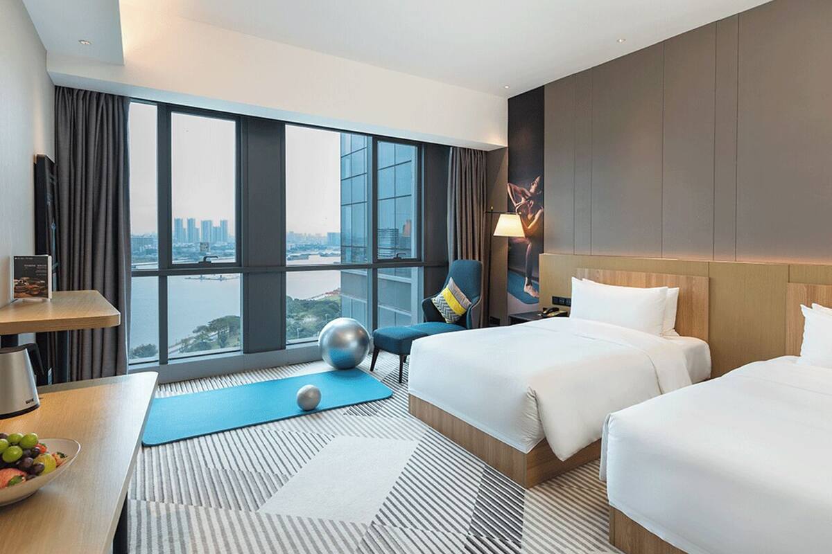 Une chambre d’hôtel avec deux lits, un bureau sur lequel se trouve un panier de fruits, un tapis de yoga et une grande fenêtre donnant sur la ville.