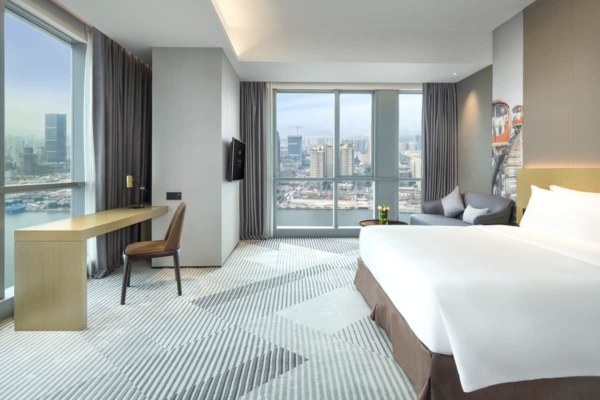 Une chambre d’hôtel moderne dotée d’un grand lit, d’un bureau et d’une chaise. Elle offre une vue sur la ville grâce à de grandes fenêtres.