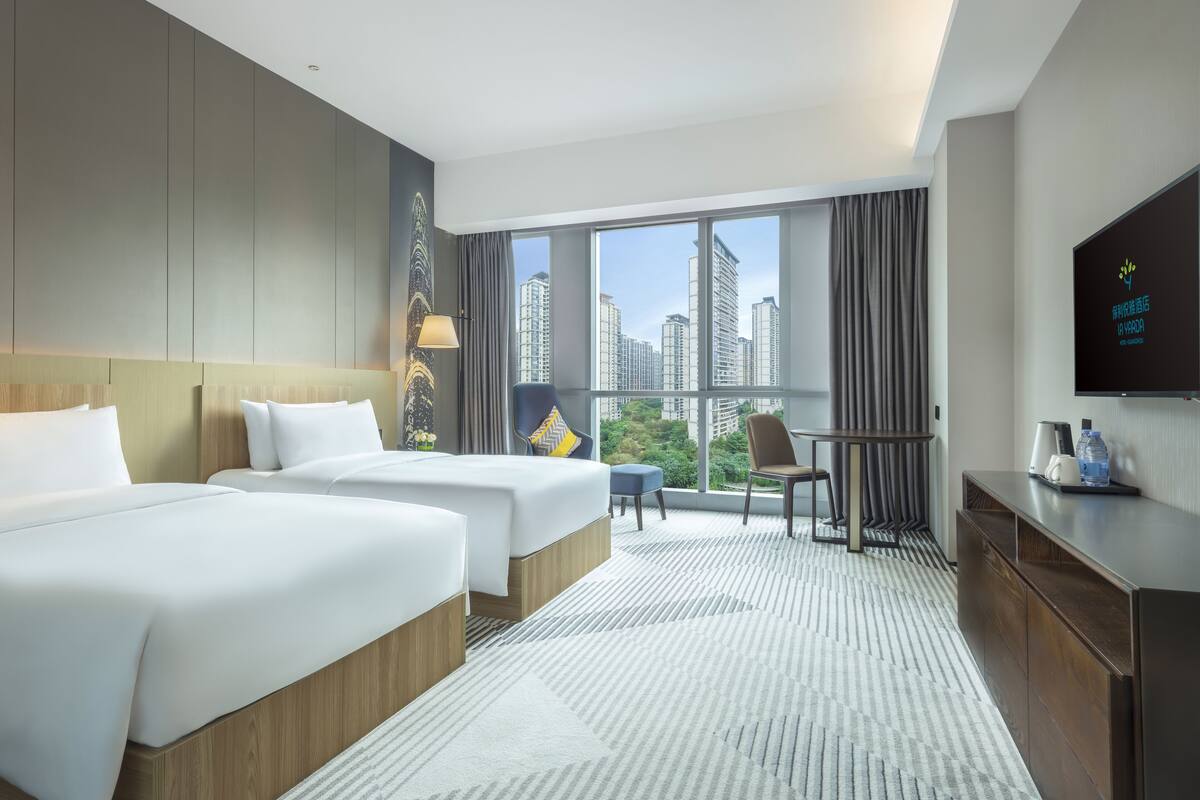 Une chambre d’hôtel avec deux lits, un bureau, une chaise, une télévision et une grande fenêtre donnant sur la ville.