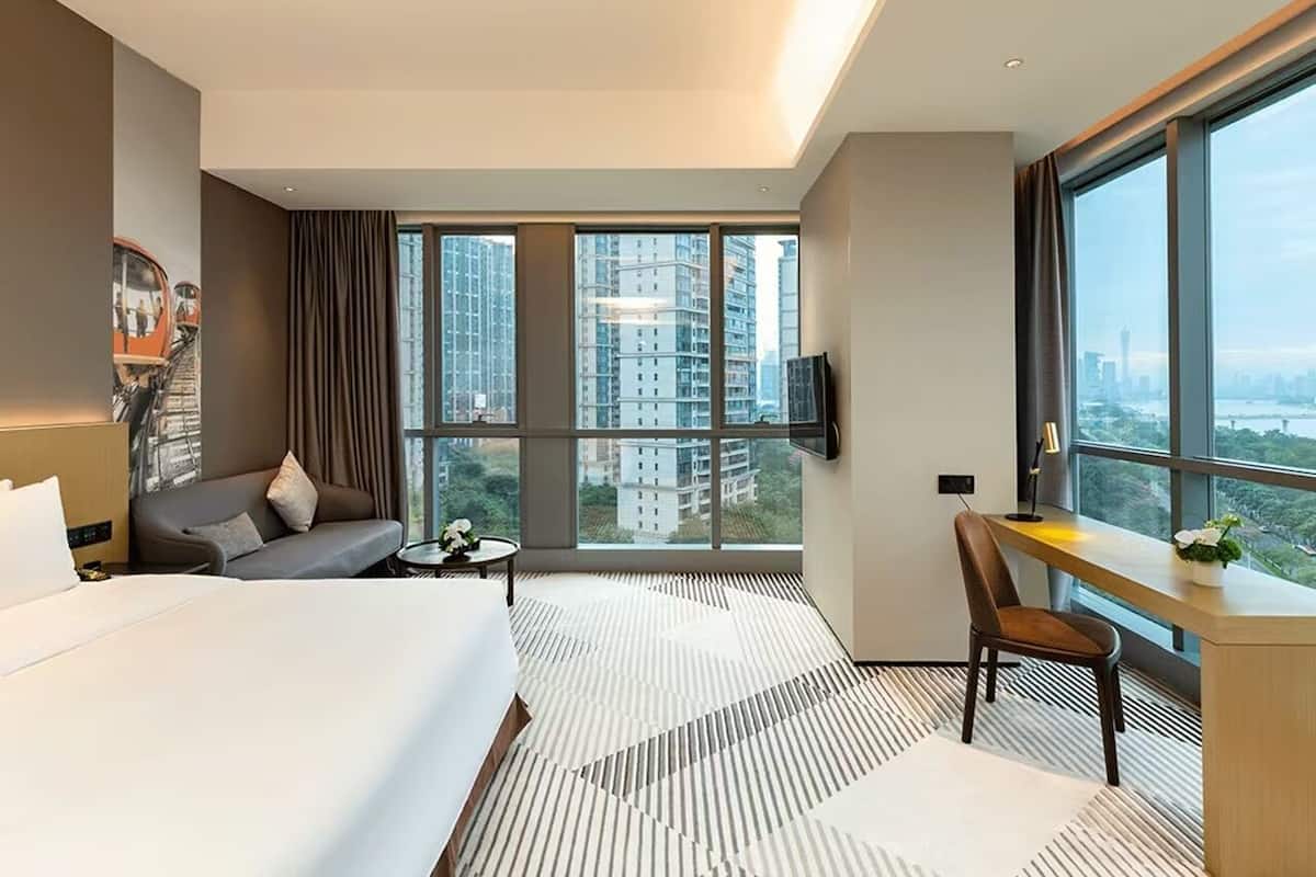 Une chambre d’hôtel moderne dotée d’un grand lit, d’un canapé, d’un bureau et offrant une vue sur les grands immeubles à travers de larges fenêtres.