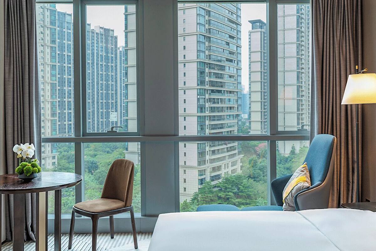 Une chambre d’hôtel dotée d’une grande fenêtre donnant sur la ville, d’un lit, de deux chaises, d’une petite table avec un vase de fleurs et d’une lampe.