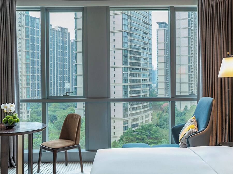 A hotel room with a large window offering a city view, a bed, two chairs, a small table with a flower vase, and a lamp.