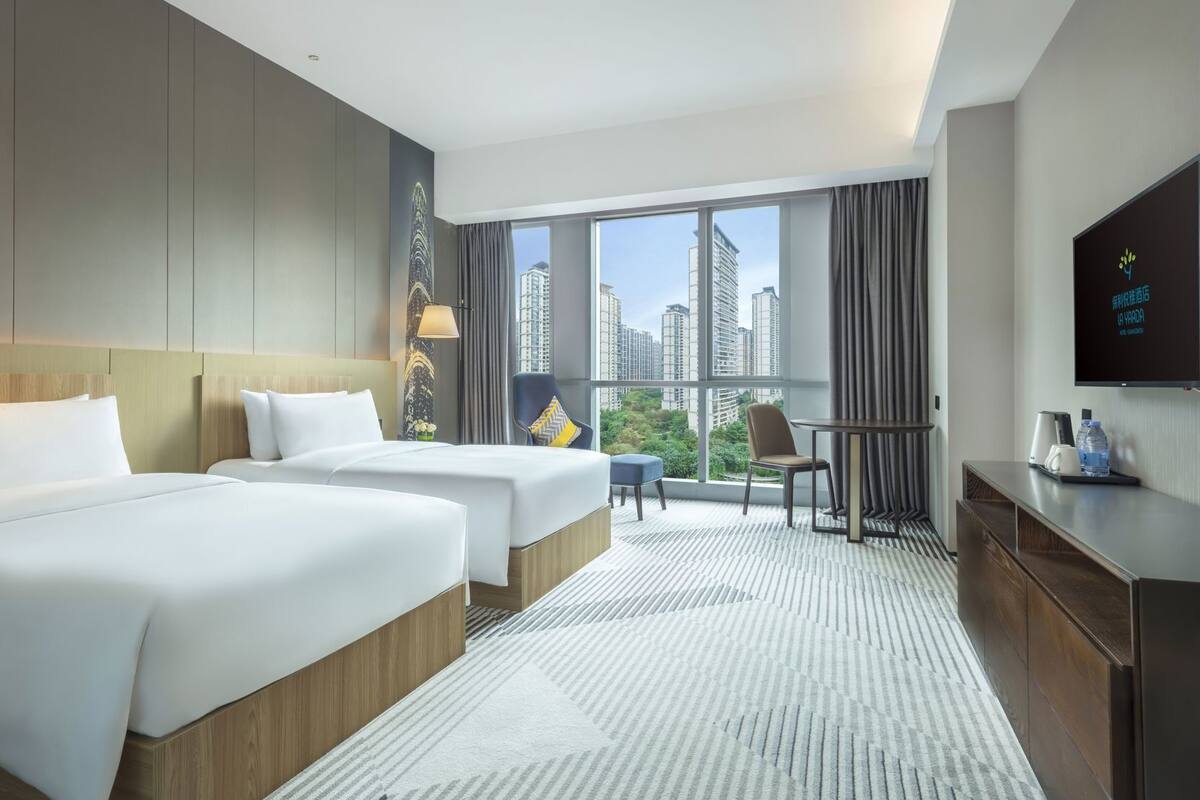 Une chambre d’hôtel avec deux lits, un bureau, une chaise, une télévision et une grande fenêtre donnant sur la ville.
