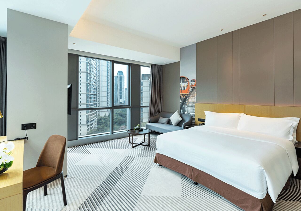 A modern hotel room with a large bed, a desk, a chair, and a view of the city through large windows.