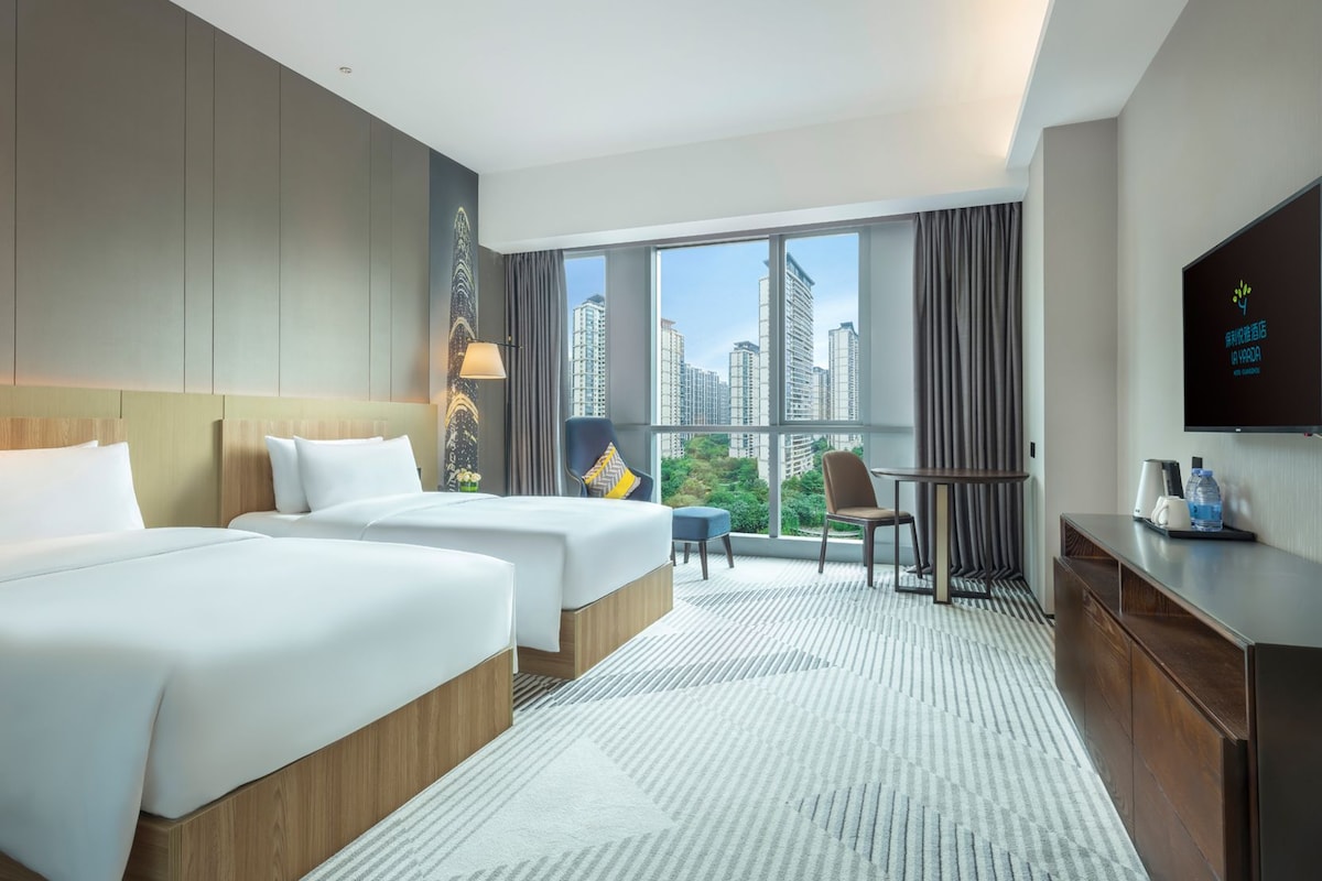 Habitación de hotel con dos camas, un escritorio, una silla, un televisor y un ventanal con vistas a la ciudad.