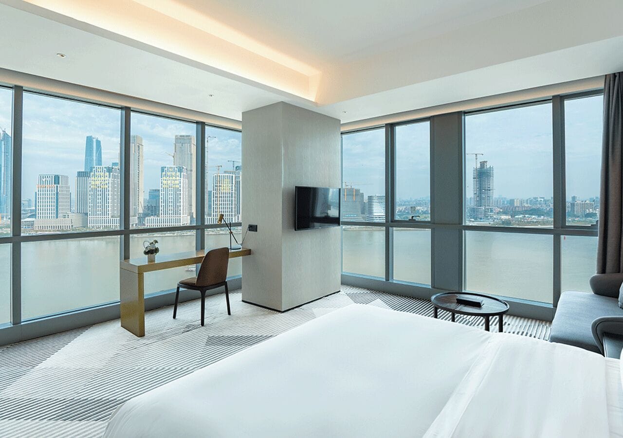 Une chambre d’hôtel moderne dotée d’un grand lit, d’un bureau avec une chaise, d’une télévision et offrant une vue sur les toits de la ville grâce à de grandes fenêtres.