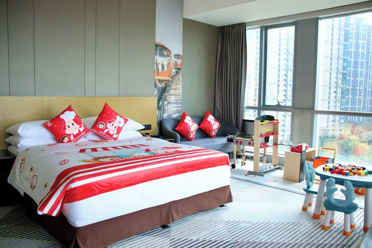 Habitación de hotel con una cama grande, un escritorio con computadora, un ventanal con vista a la ciudad y una zona de juegos para niños.