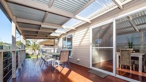 Une terrasse couverte avec un plancher en bois, des meubles d’extérieur et une porte-fenêtre coulissante donnant sur un espace intérieur aménagé avec des meubles en osier.
