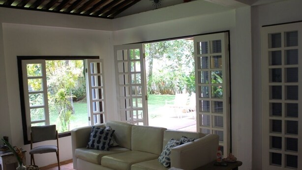 A room with a beige sofa, a wooden table, and a chair. There are large windows with white frames and a view of greenery outside.