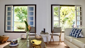 A living room with a glass coffee table, two chairs, a sofa, and a view of greenery through open windows.