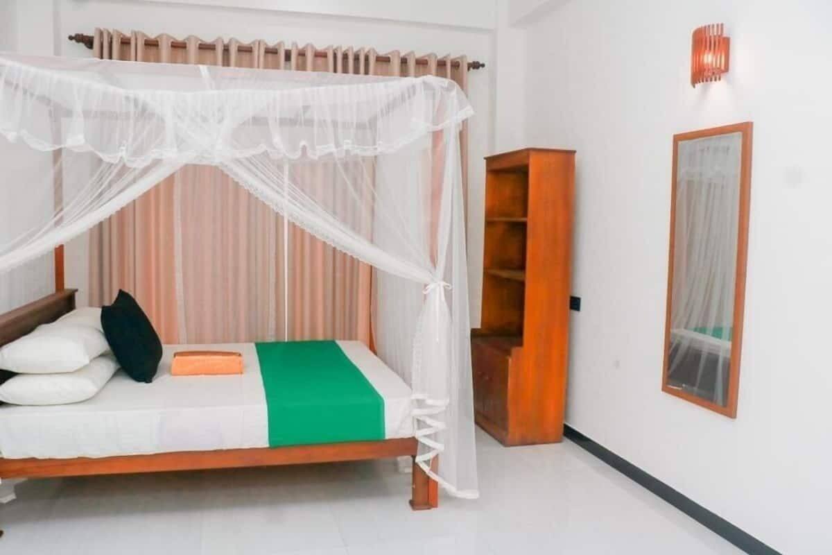 A single bed with a canopy, a wooden nightstand, a green bedspread, and a small wooden cabinet against a white wall.