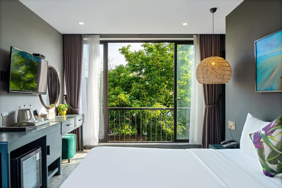Une chambre d’hôtel moderne dotée d’un grand lit, d’un bureau avec un téléviseur à écran plat, d’un minibar et d’un balcon offrant une vue sur les arbres.
