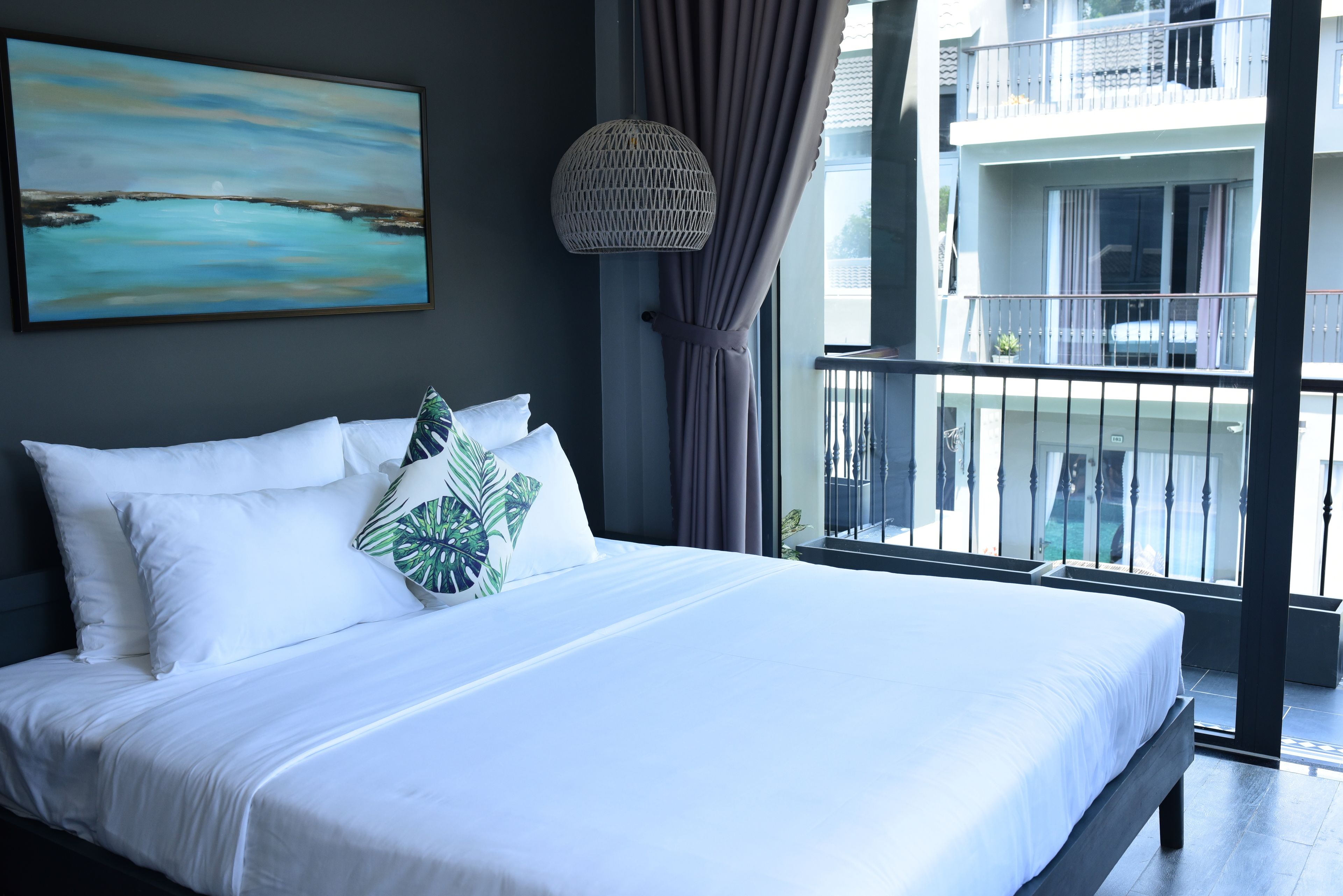 A neatly made bed with white linens and a tropical leaf pillow, a large painting of a coastal scene, and a balcony with a wrought iron railing and a hanging light fixture.