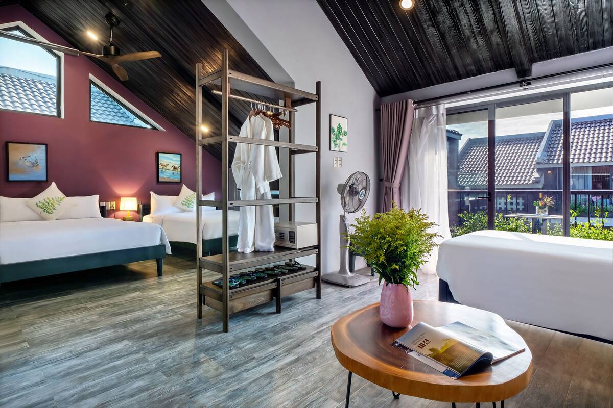 Une chambre d’hôtel moderne avec un lit, une étagère en bois où sont posés des vêtements, un ventilateur, une table avec un magazine et une fenêtre donnant sur des immeubles.