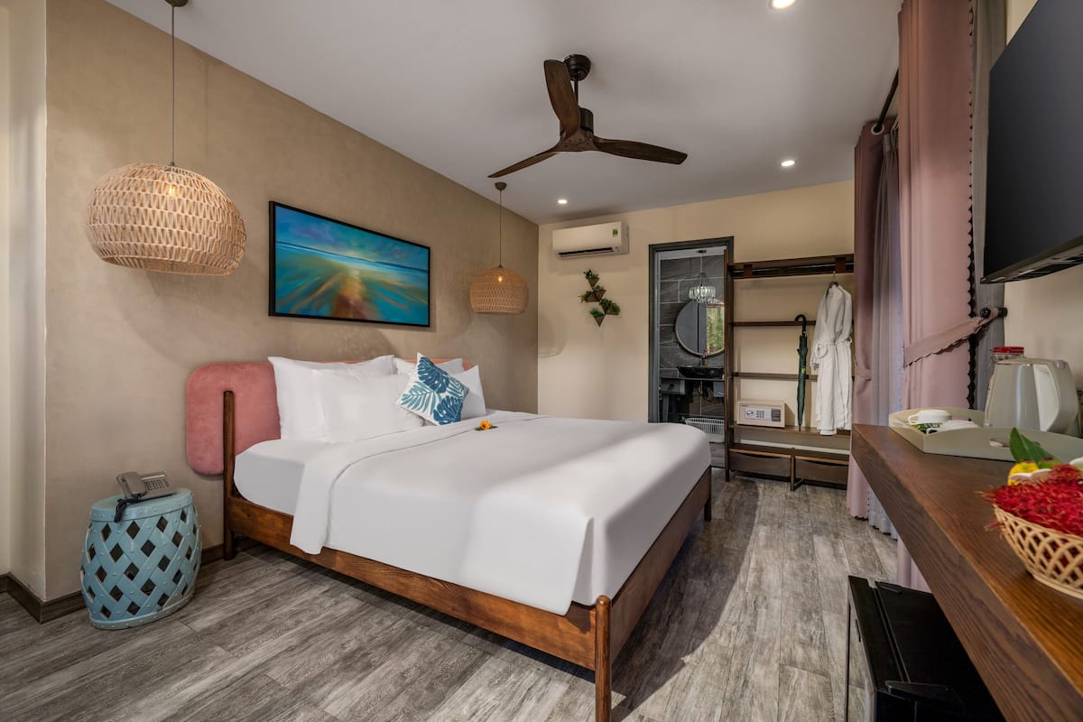 Une chambre d’hôtel moderne avec un lit en bois, un ventilateur de plafond, une grande fenêtre et une salle de bain visible à travers une porte ouverte.