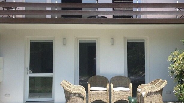 Een overdekt terras met rieten stoelen en een tafel, een schuifdeur en een balkon met een hekwerk.