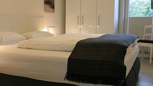 Een slaapkamer met een bed, witte kussens, een zwarte plaid, een nachtkastje en een raam met jaloezieën.