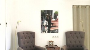 Een kamer met twee grijze fauteuils, een klein tafeltje met een klok en een ingelijst schilderij aan de muur.