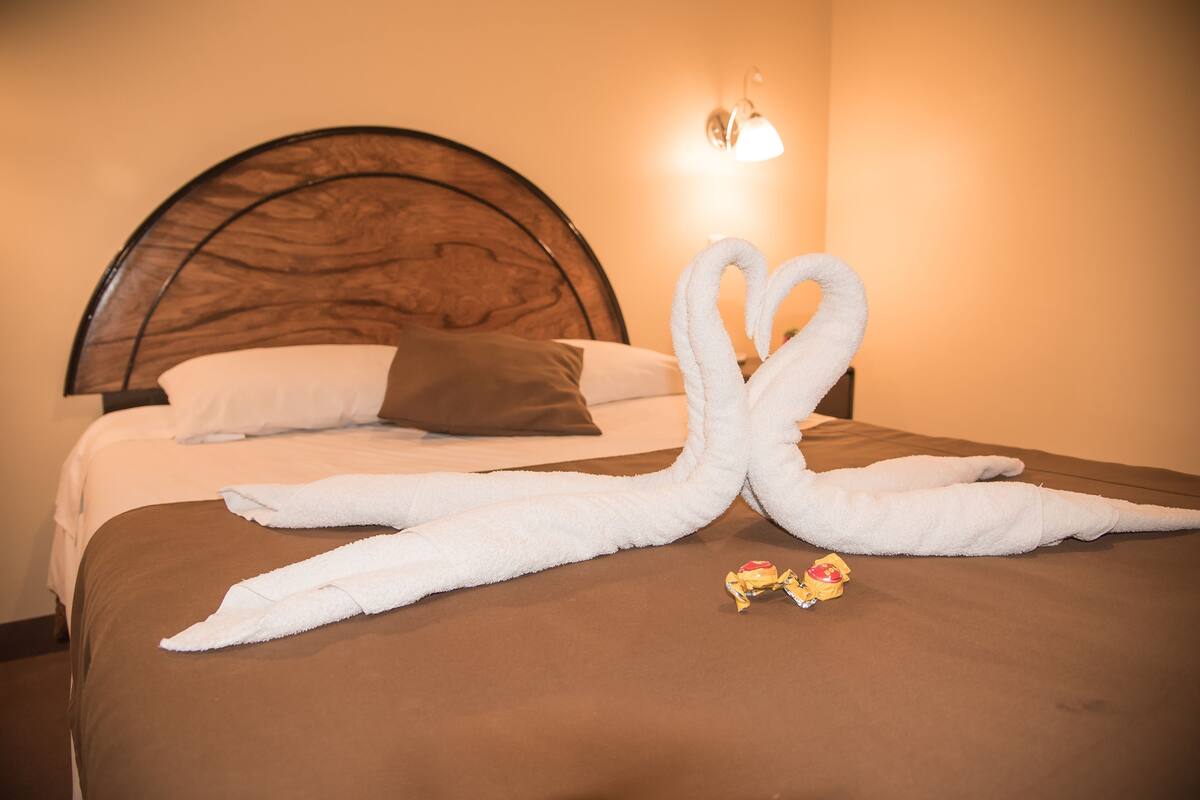 A hotel room with a bed, a brown headboard, a wall-mounted light fixture, and two towels arranged in a heart shape.