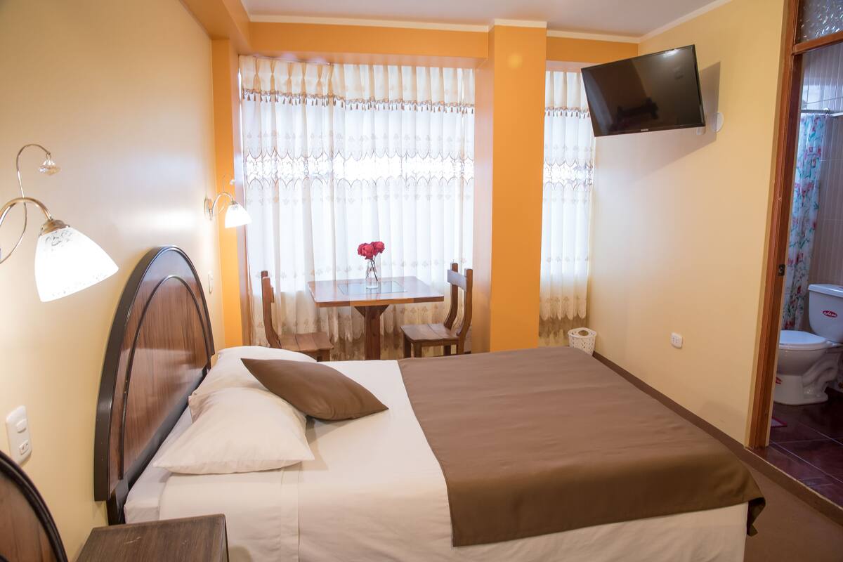 A hotel room with a bed, a desk, a chair, a TV mounted on the wall, and a bathroom visible through an open door.