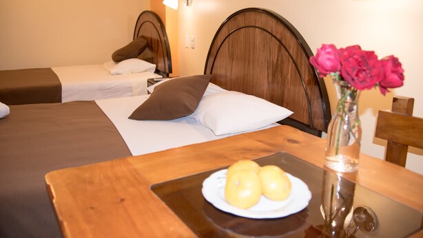 A hotel room with two beds, a wooden table with a plate of fruit, and a vase with flowers. - Samary Hostel (Puno)