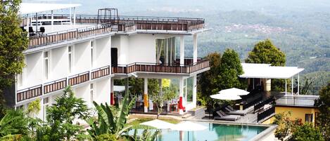 A modern two-story building with a swimming pool, outdoor seating, and a view of the surrounding landscape.