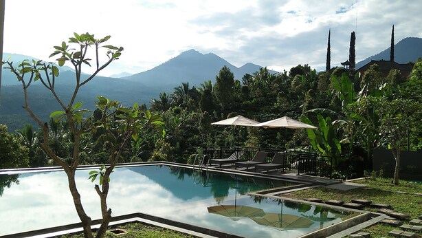 A pool with a clear view of mountains in the background, surrounded by lush greenery and a traditional building with a peaked roof. - Atres Sari Resort (Munduk)