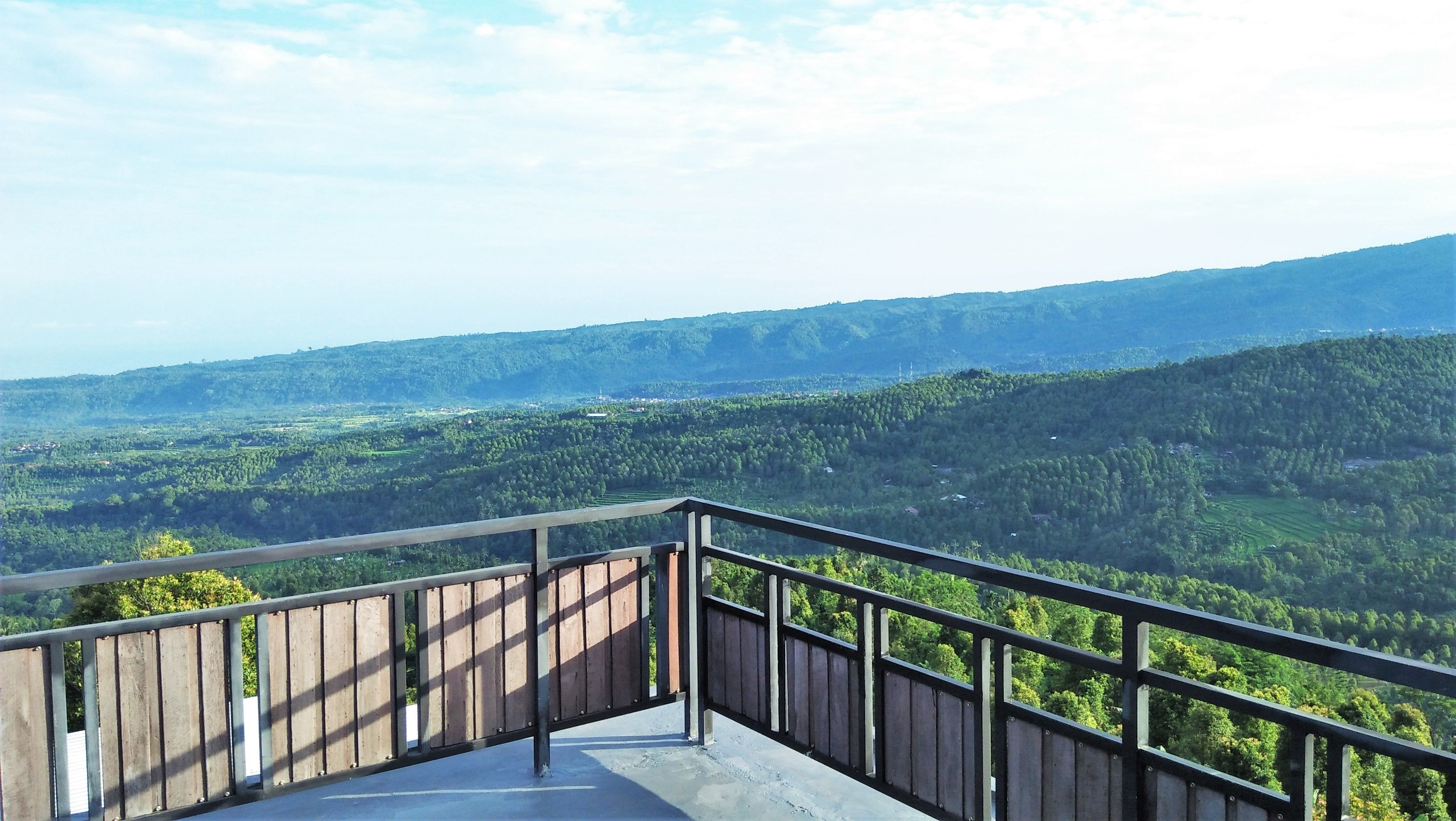 Ein Balkon mit Metallgeländer, der auf eine gebirgige Landschaft mit dichtem Grün und blauem Himmel blickt.