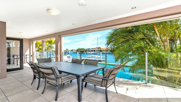 A modern dining area with a large table and chairs, overlooking a marina with boats and palm trees.