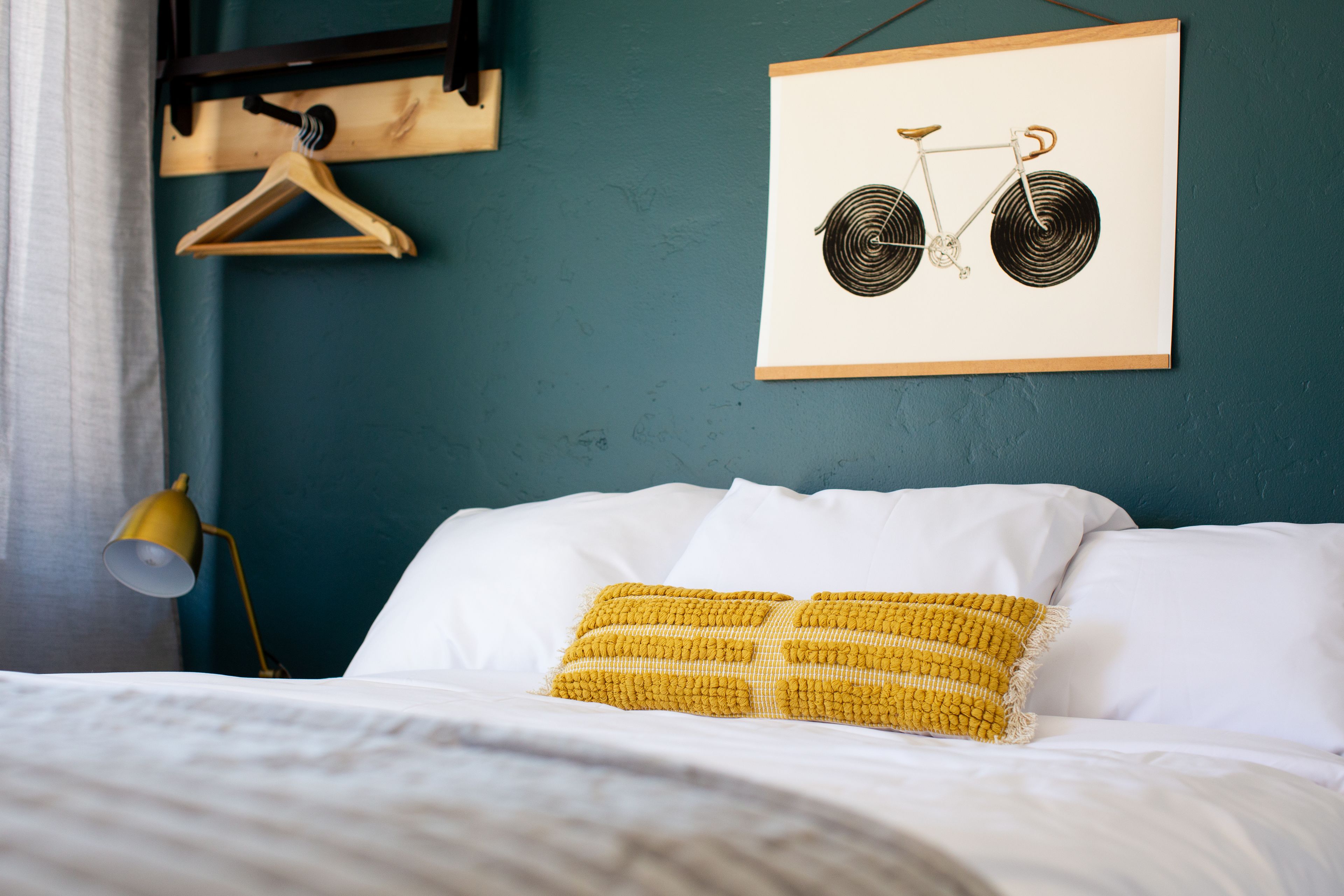 Una cama bien hecha con ropa blanca y un cojín amarillo de punto. Encima de la cama, en la pared verde azulada, cuelga una obra de arte en blanco y negro de una bicicleta enmarcada.