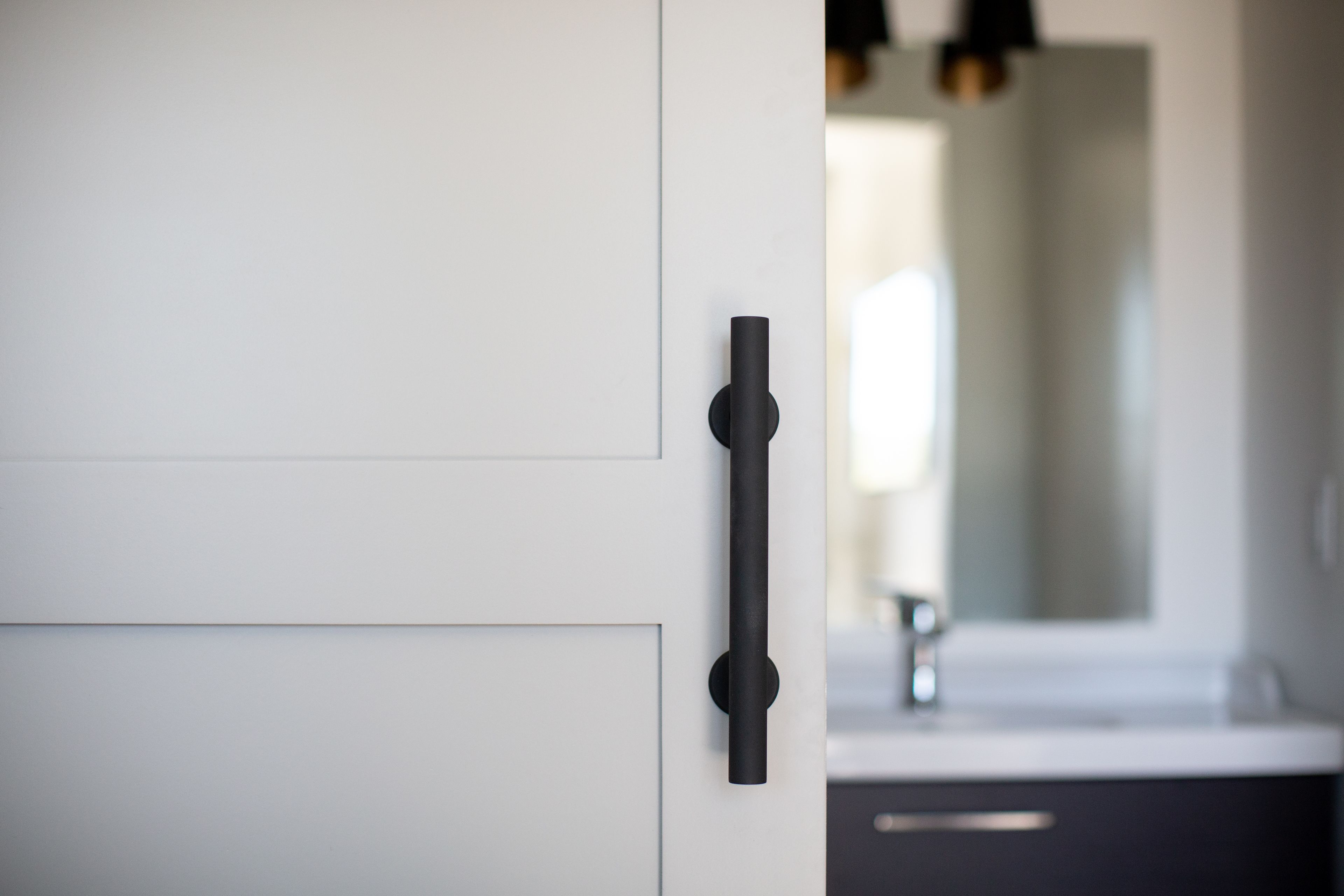 A modern bathroom with a minimalist design, featuring a white door with a black handle, a dark cabinet, and a large mirror above a sink.