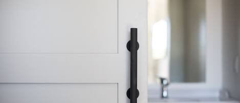Un baño moderno con un diseño minimalista, que incluye una puerta blanca con pomo negro, un mueble oscuro y un espejo grande sobre un lavamanos.