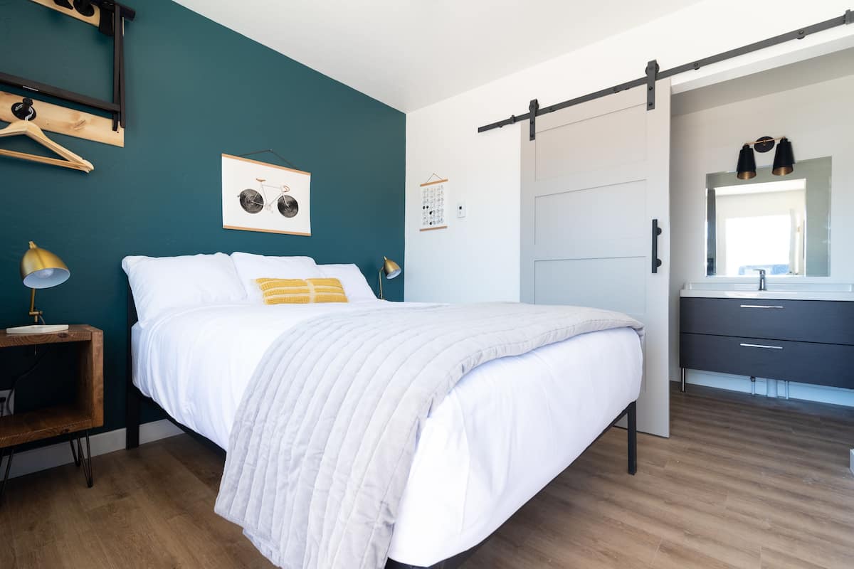 Une chambre à coucher comprenant un lit, une table de chevet, une étagère fixée au mur, une porte et une salle de bain avec un miroir et un lavabo.