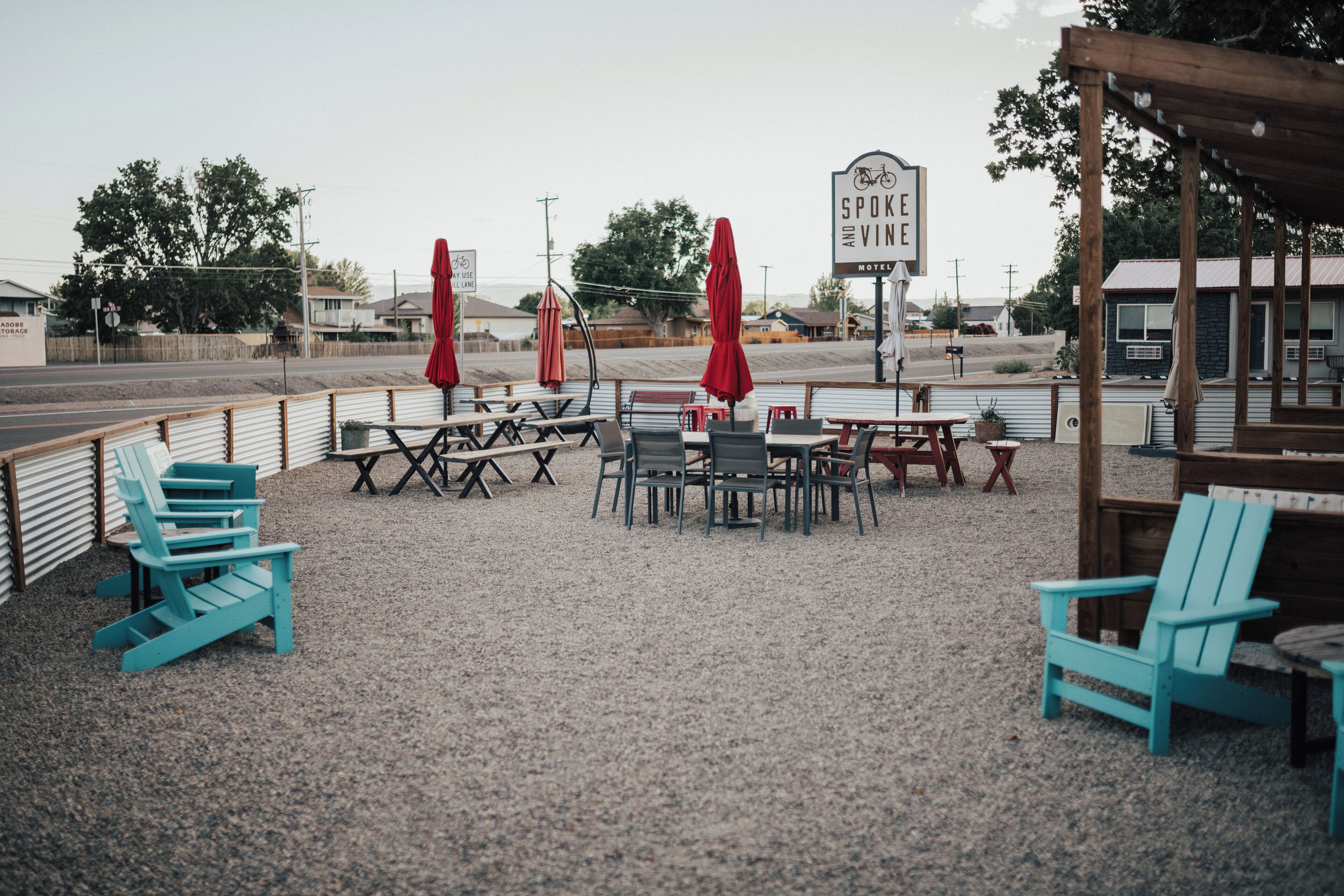 A gravel patio with outdoor seating, including Adirondack chairs and tables, under a covered area with red umbrellas. A sign for the Spokane Ave Motel is visible.