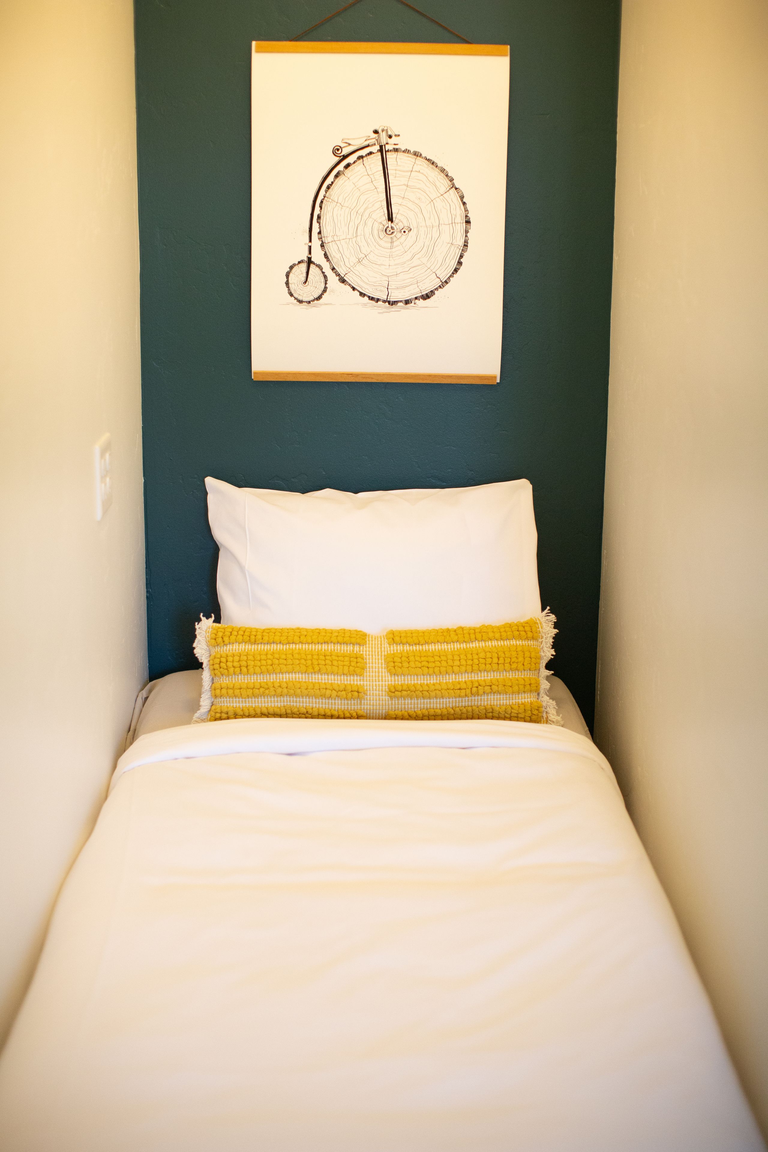 A bed with white bedding and a yellow woven throw pillow, a white pillow, and a wall-mounted clock.