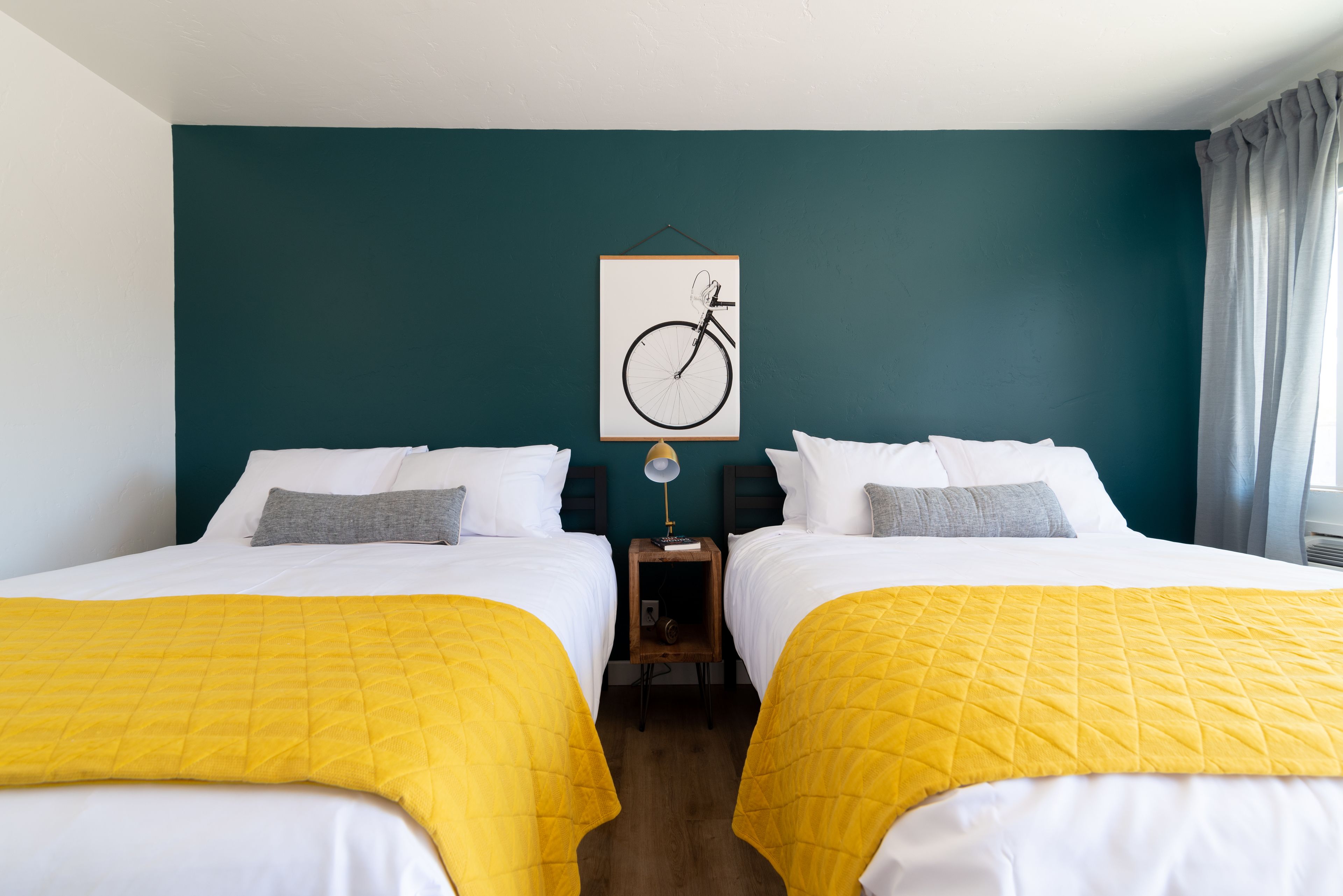 A hotel room with two beds, each with a yellow quilted bedspread, white bedding, and gray pillows. A framed bicycle artwork hangs on the teal-colored wall above the beds.