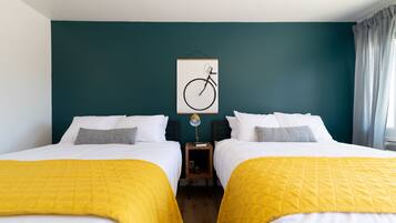 A hotel room with two beds, each with a yellow quilted bedspread, white bedding, and gray pillows. A framed bicycle artwork hangs on the teal-colored wall above the beds.