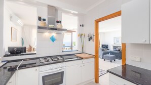 Een moderne keuken met witte kasten, een zwart aanrecht en een kookplaat. Er is een raam met gordijnen, een houten deur en uitzicht op een woonkamer met een bank en een tafel.