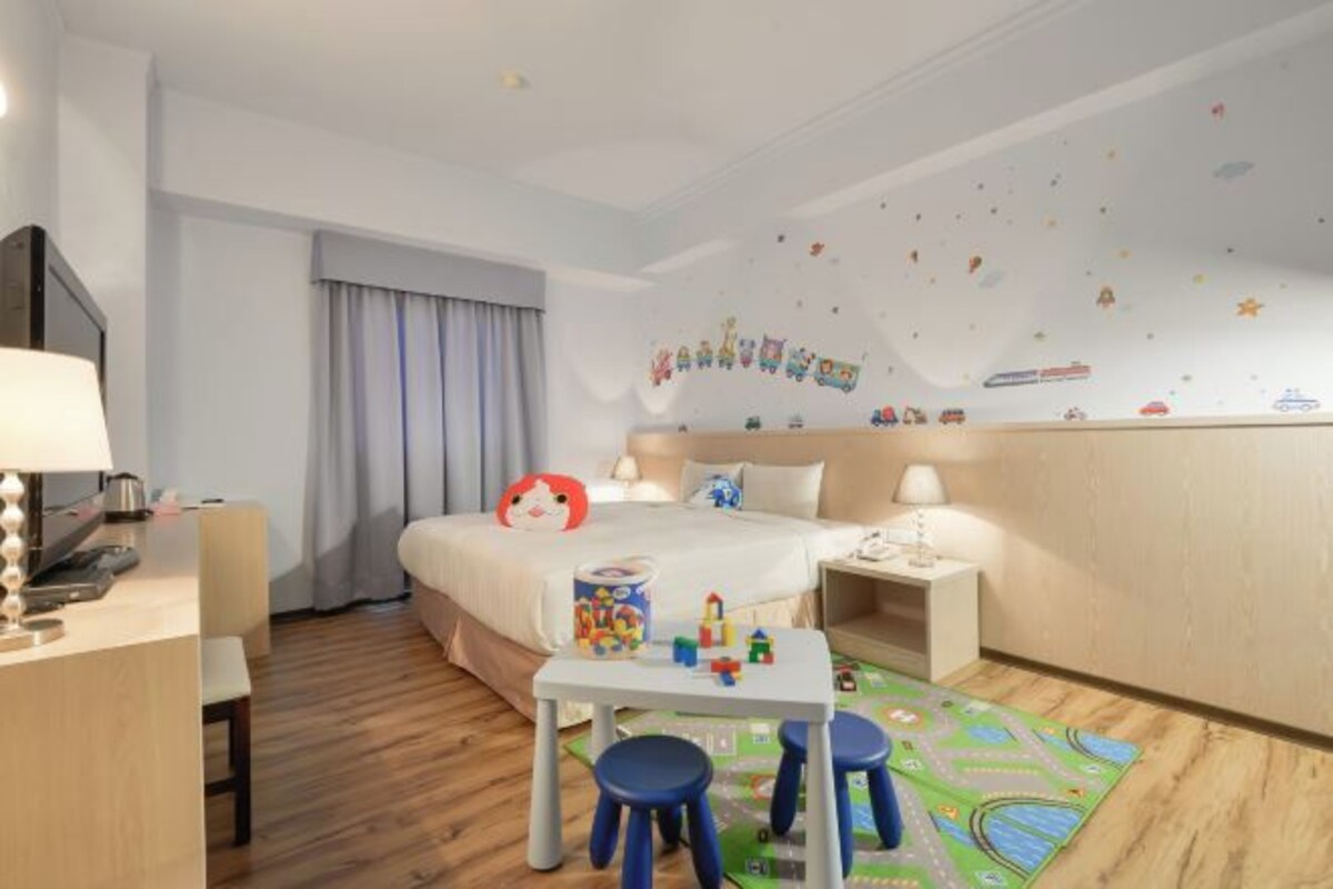 A hotel room with a bed, bedside table, lamp, desk, chair, and a play area with toys and a mat.