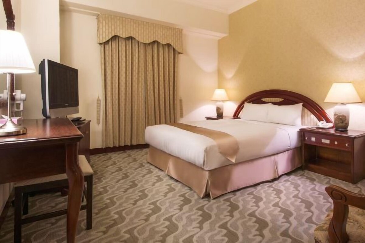 A hotel room with a bed, bedside lamps, a desk with a lamp, a television, and a curtain.