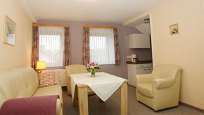 A room with a sofa, a table with a vase of flowers, a chair, and a kitchenette in the background. - Ferienhof Jens Apartment 4 - Apartment 4 (Kronsgaard)