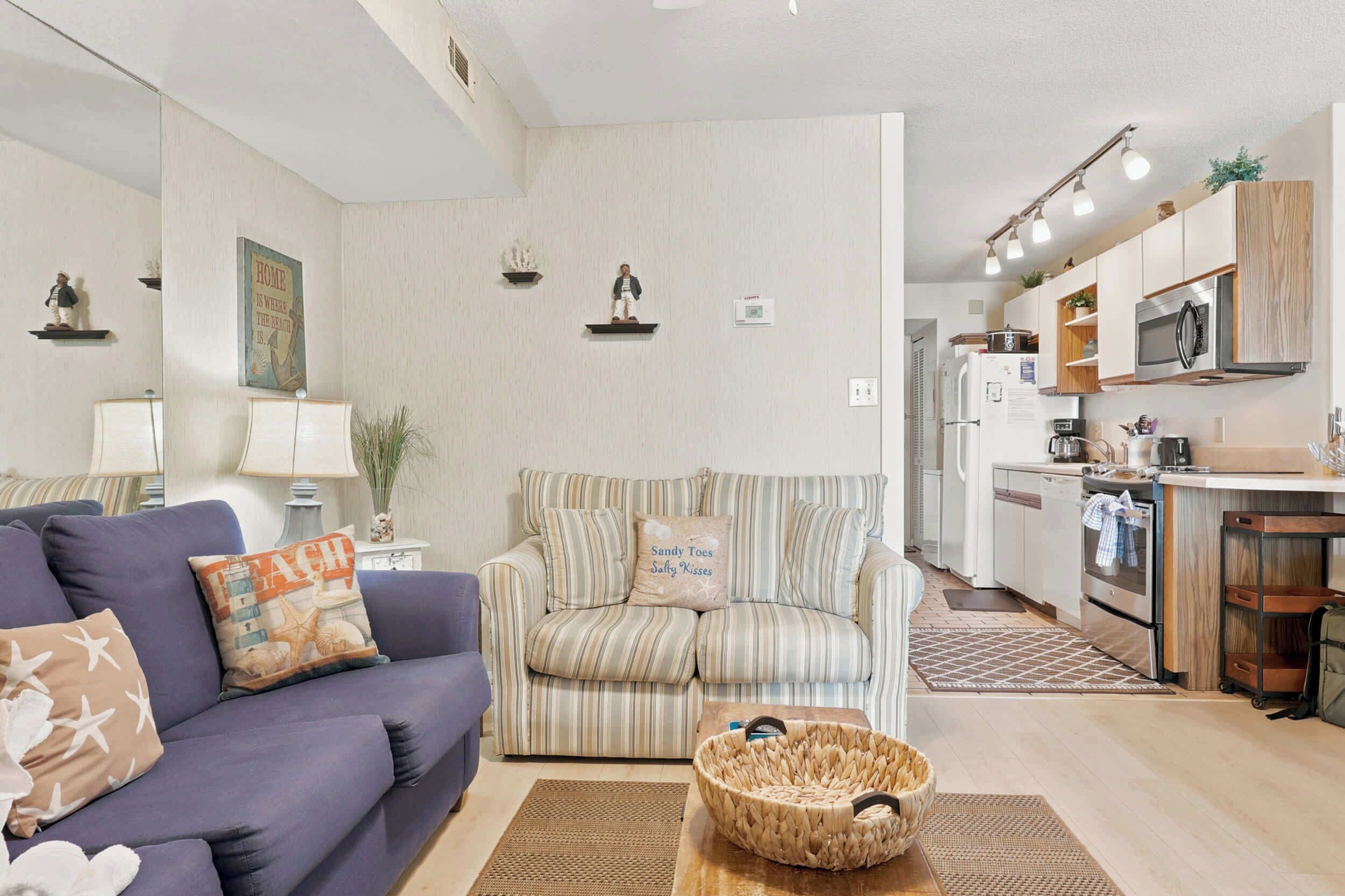 Een woonkamer met een bank, een gestreept fauteuil, een gevlochten mand en een keukenhoek met moderne apparatuur.