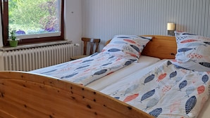 Een netjes opgemaakt bed met een beddekleed met patroon, een houten hoofdbord, een bedlampje en uitzicht op groen door een raam.