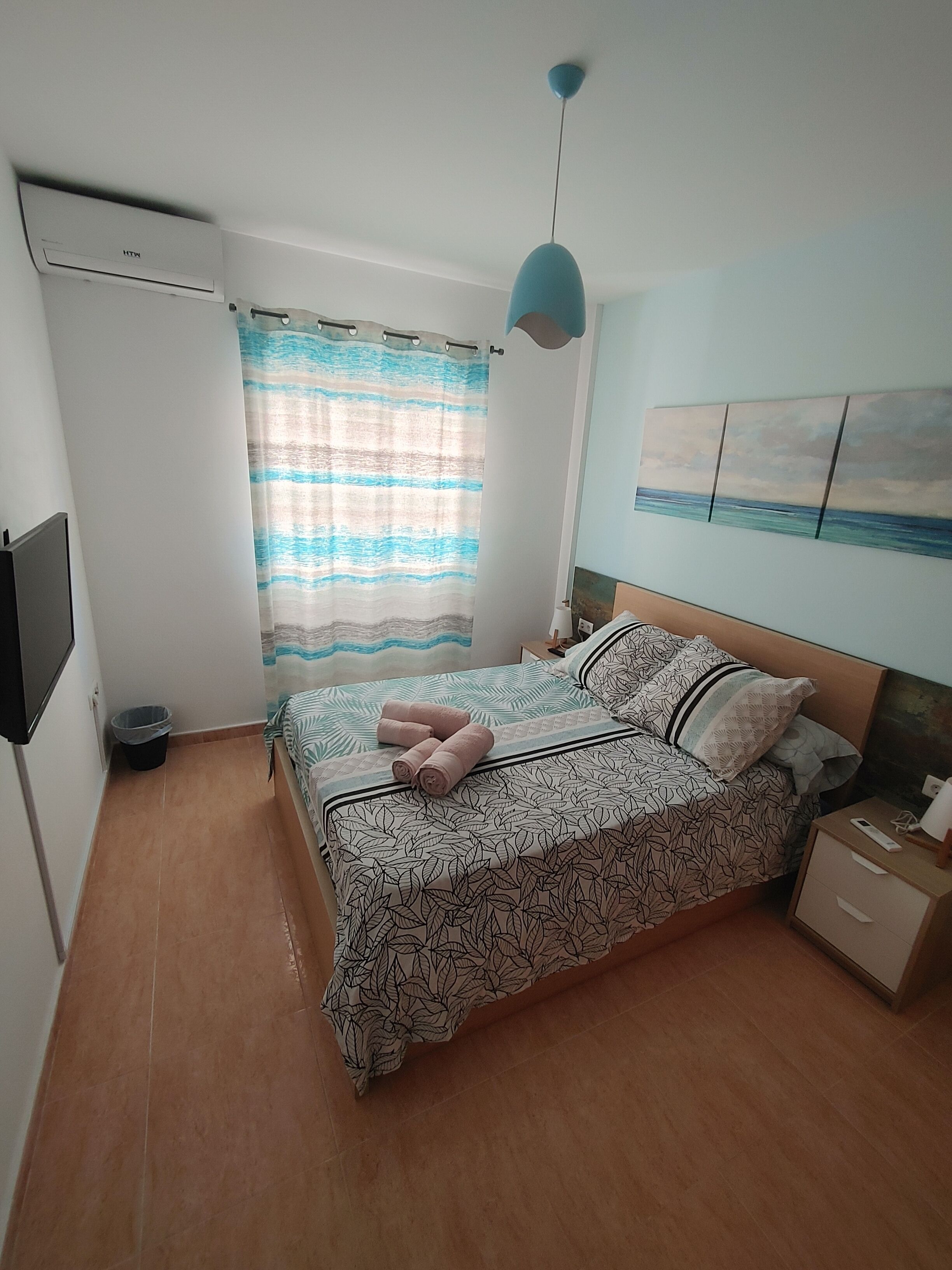 A bedroom with a bed, a nightstand, a wall-mounted TV, a window with a curtain, and a painting of a beach scene.