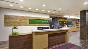 A modern hotel reception area with a wooden reception desk, a green sign for 'Home2 Suites by Hilton', and a counter with various items. - Home2 Suites by Hilton Birmingham Fultondale (Fultondale)