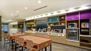 A modern restaurant with a long wooden table and chairs, a kitchen area with a microwave, and a display of food items. - Home2 Suites by Hilton Birmingham Fultondale (Fultondale)